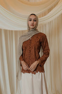 A woman wearing a rust-brown embroidered long-sleeve top and a cream maxi skirt, paired with a light beige hijab, standing in front of a draped cream fabric backdrop with floral arrangements at the base.