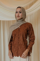 A woman wearing a rust-brown lace long-sleeve blouse with a button front and a cream maxi skirt, styled with a beige hijab. She is standing in front of a draped cream fabric backdrop with floral arrangements at the base.