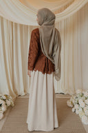 A woman wearing a brown lace long-sleeve top and a cream maxi skirt, paired with a taupe hijab, standing in a decorated indoor setting with draped cream fabric, floral arrangements, and a woven runner.